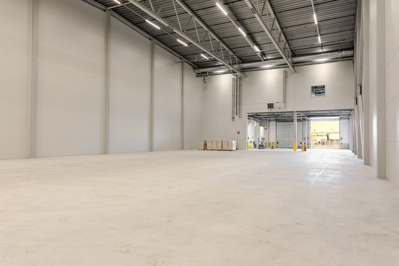 Stor, tom lagerlokal med högt i tak, slätt betonggolv, industriell belysning och några staplade lastpallar vid en lastbrygga längst bort.