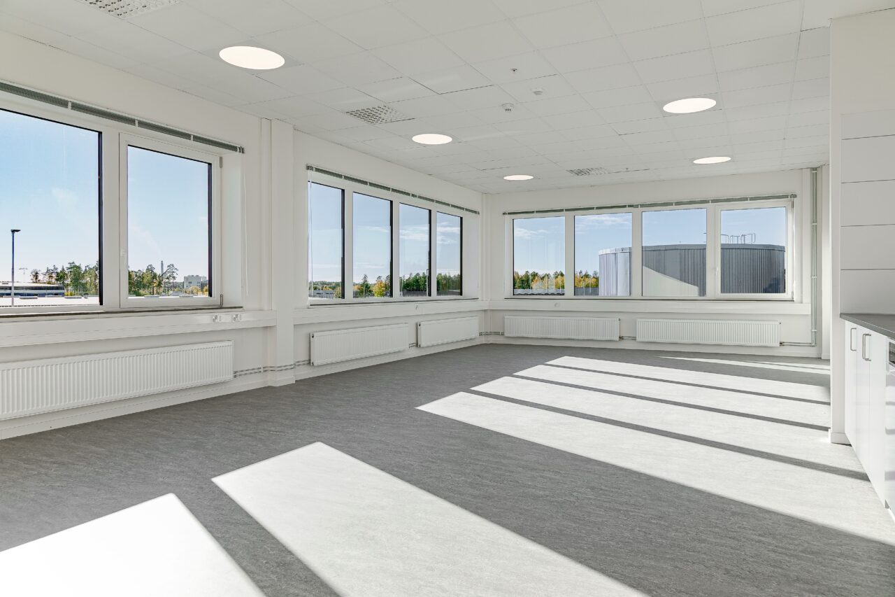 Ett stort, tomt kontorsrum med vita väggar, grått golv och flera stora fönster som släpper in starkt solljus.