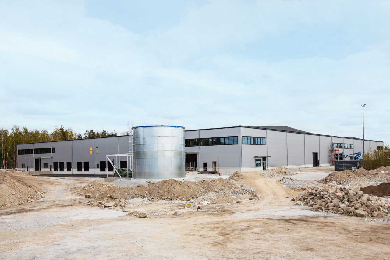 En stor, modern industribyggnad med grå fasadbeklädnad, svarta fönster och en cylindrisk metalltank framför, omgiven av jord och byggmaterial.