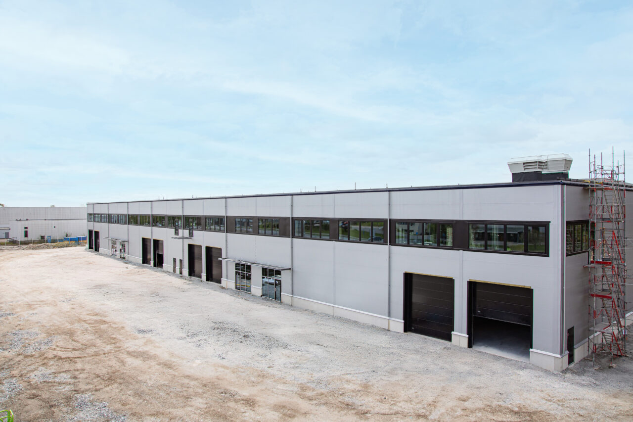 En modern, rektangulär industribyggnad under uppförande med stora fönster, flera garageportar och byggnadsställningar i ena hörnet. Den omgivande marken är asfalterad.