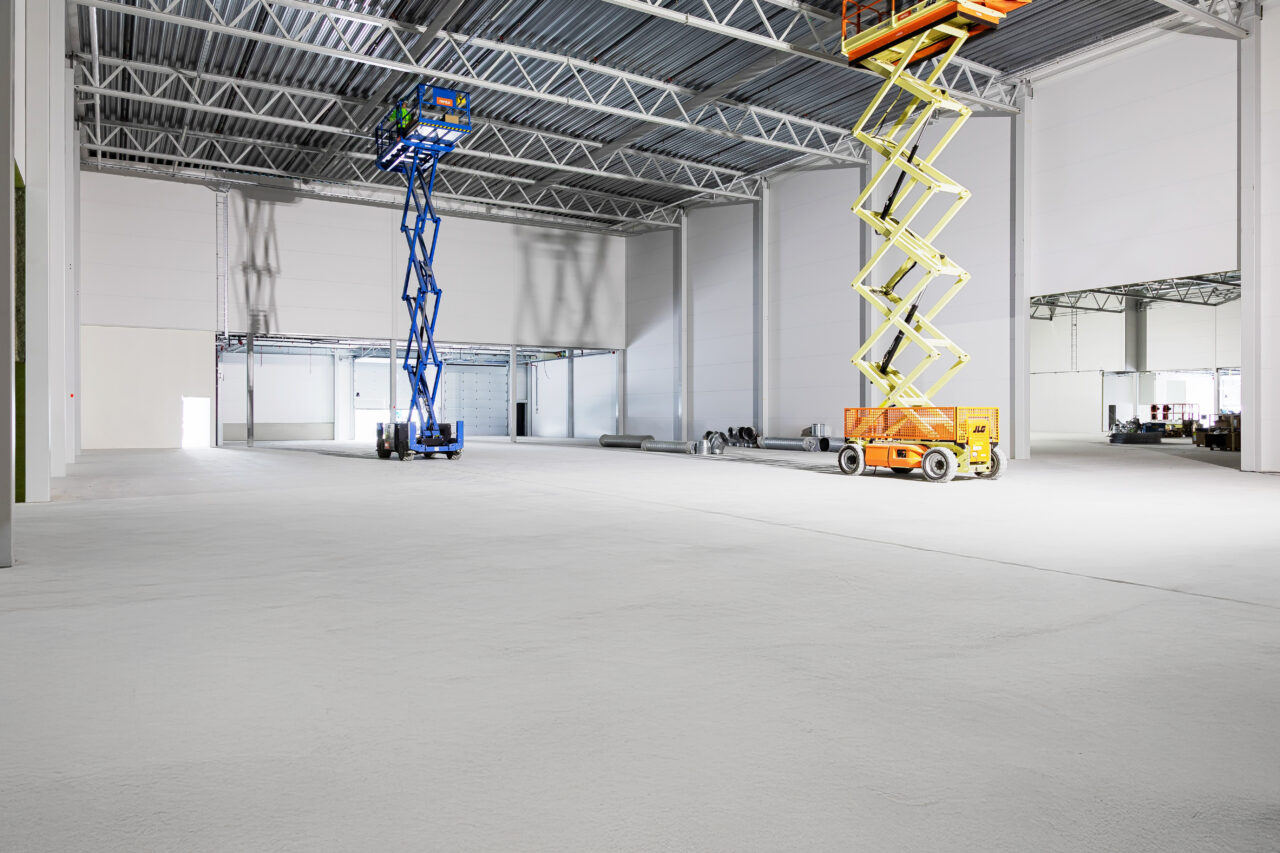 Stort tomt industrilager med högt i tak, med två mobila saxliftar - en gul och en blå - och lite byggmaterial i bakgrunden.