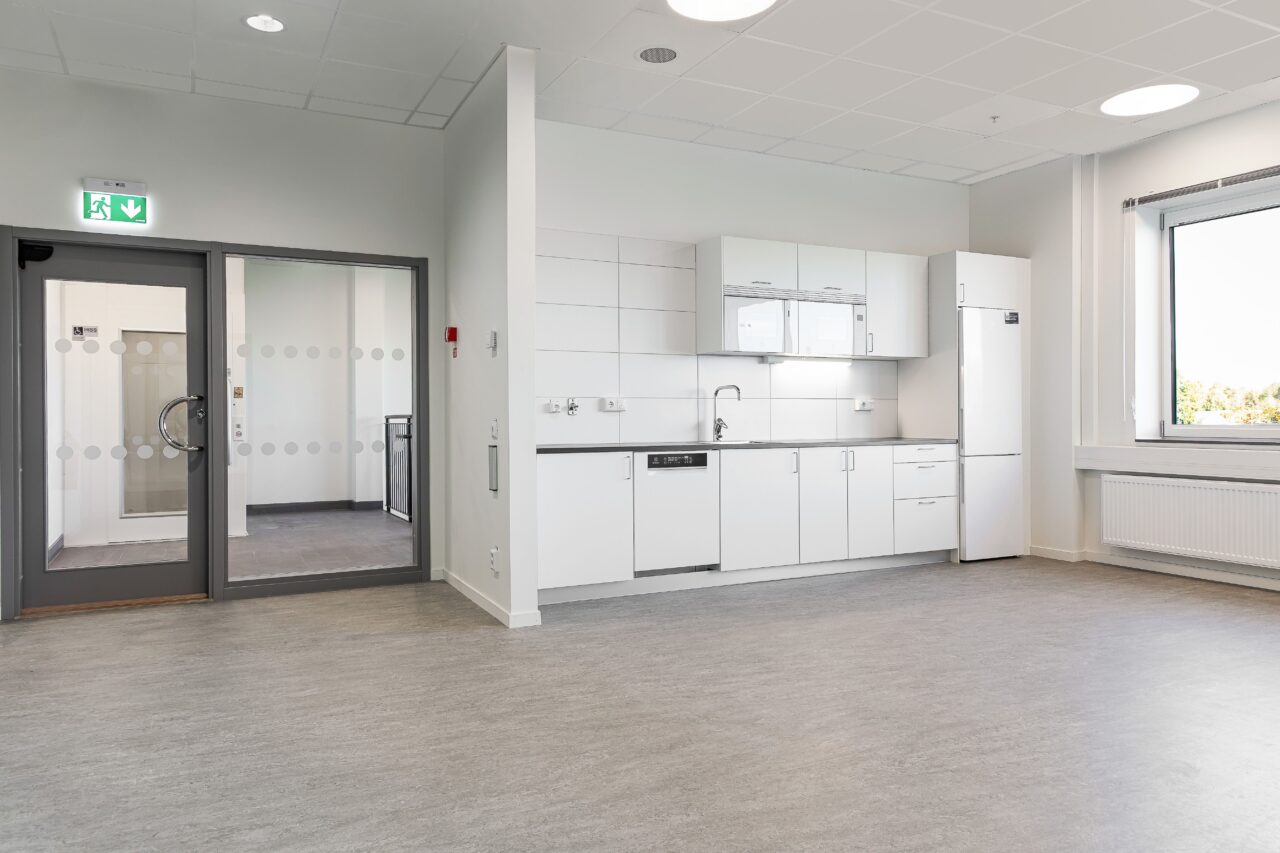 Ett modernt, tomt rum med ett litet vitt kök, grått golv, ett stort fönster och dubbla glasdörrar.