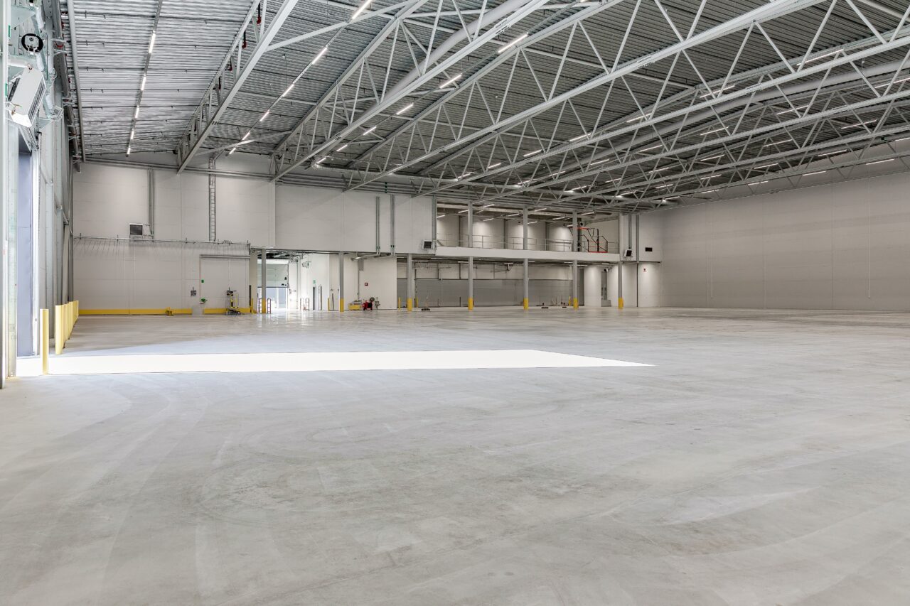 Stort, tomt industrilager med betonggolv, högt metalltak och stor öppen entré som syns på vänster sida.