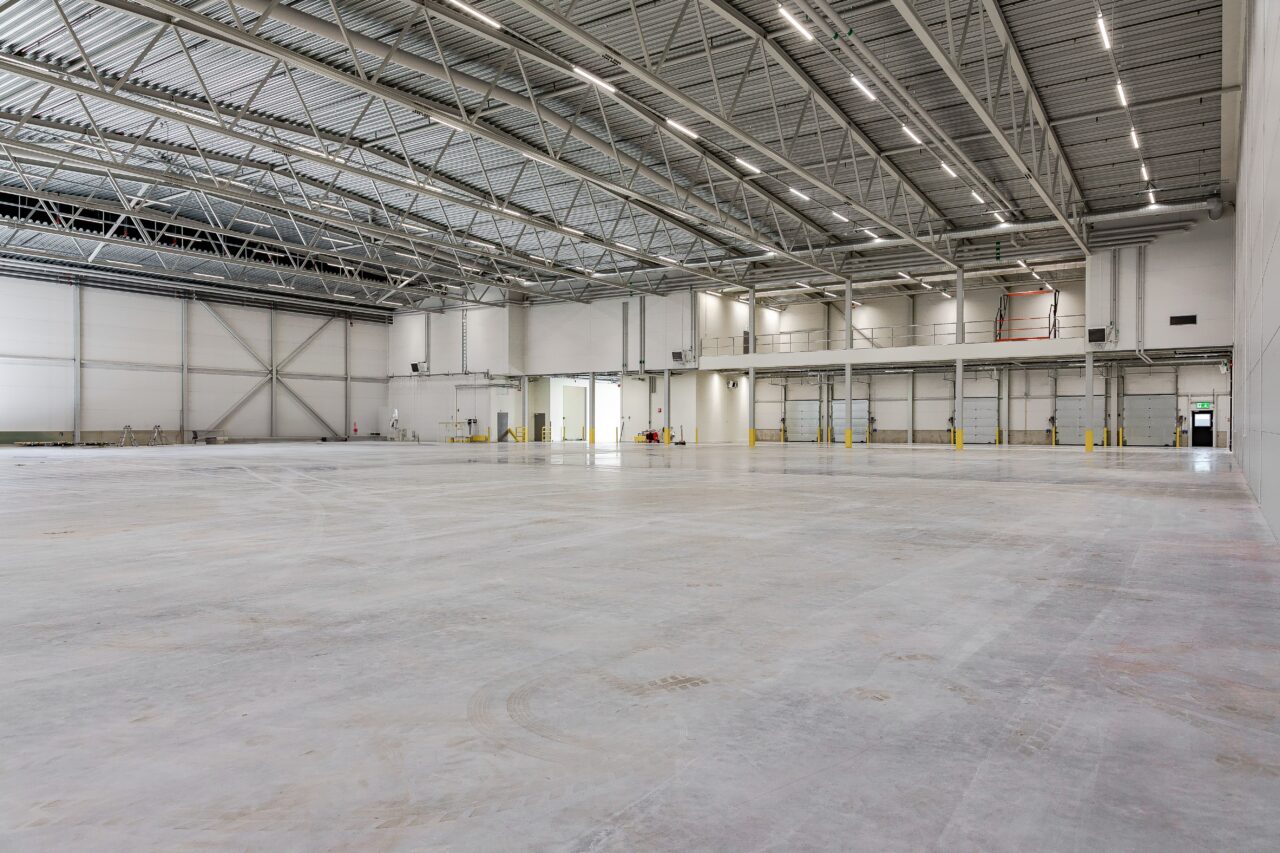 Ett tomt, rymligt industrilager med högt i tak, betonggolv och stark takbelysning. Några dörrar och ett litet upphöjt kontorsområde syns på den bortre väggen.
