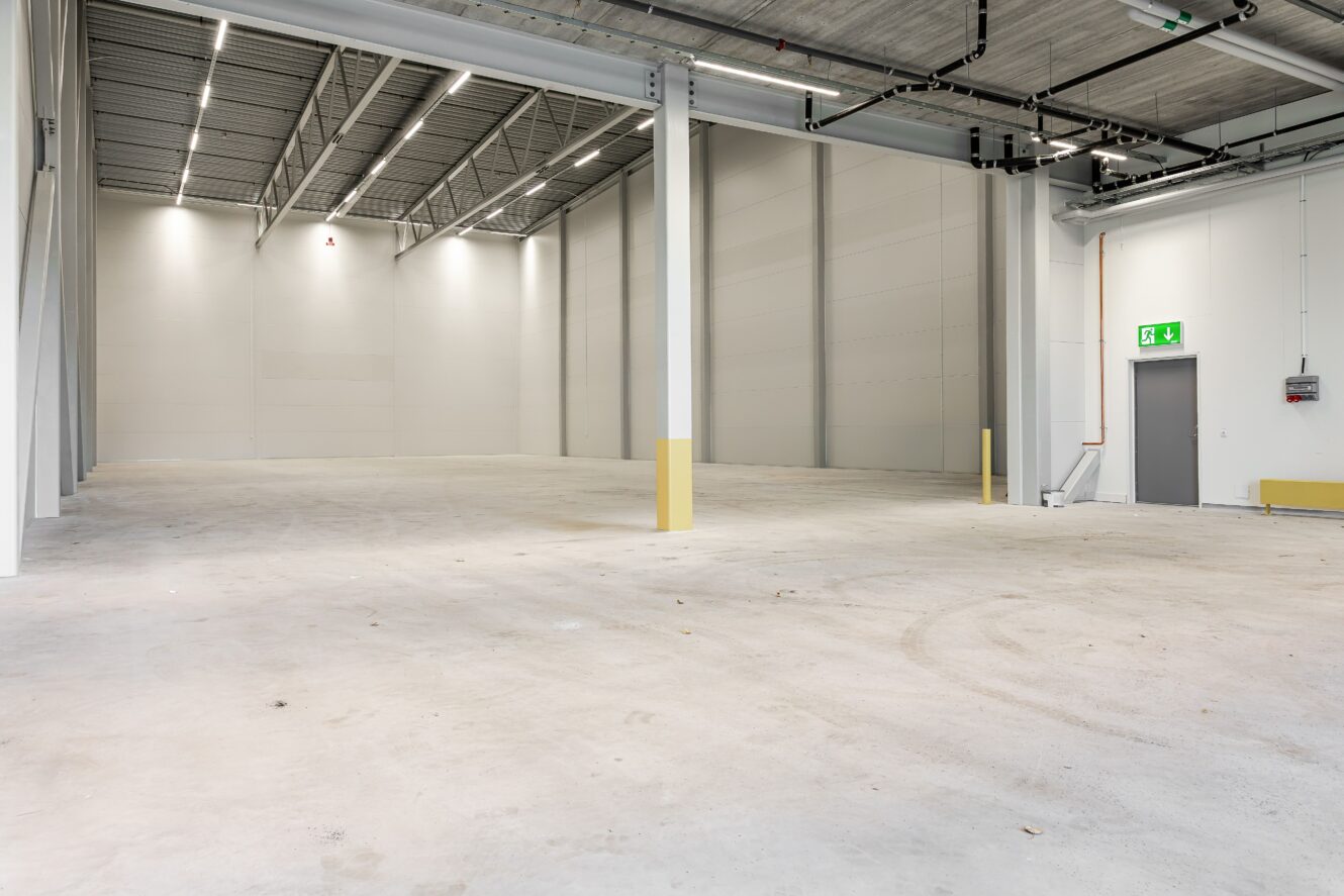 Rymlig, tom industrihall med betonggolv, högt i tak, stödpelare och en grå utgångsdörr med grön nödutgångsskylt.