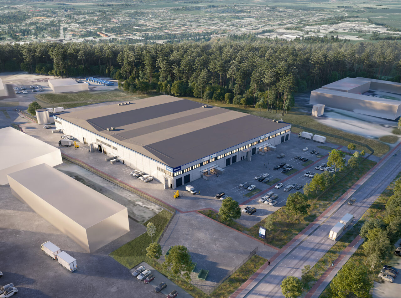 Flygfoto över en stor lagerbyggnad med parkerade fordon och lastbilar, omgiven av träd och ytterligare byggnader, intill en väg.