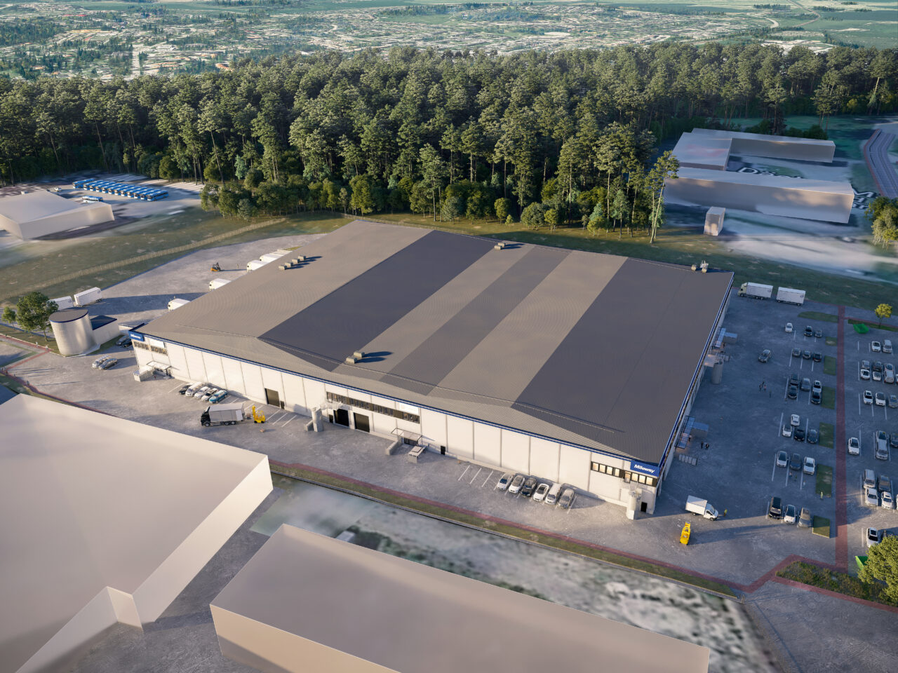 Flygfoto över en stor lagerbyggnad med parkeringsplatser och flera fordon, omgiven av träd och andra industribyggnader.