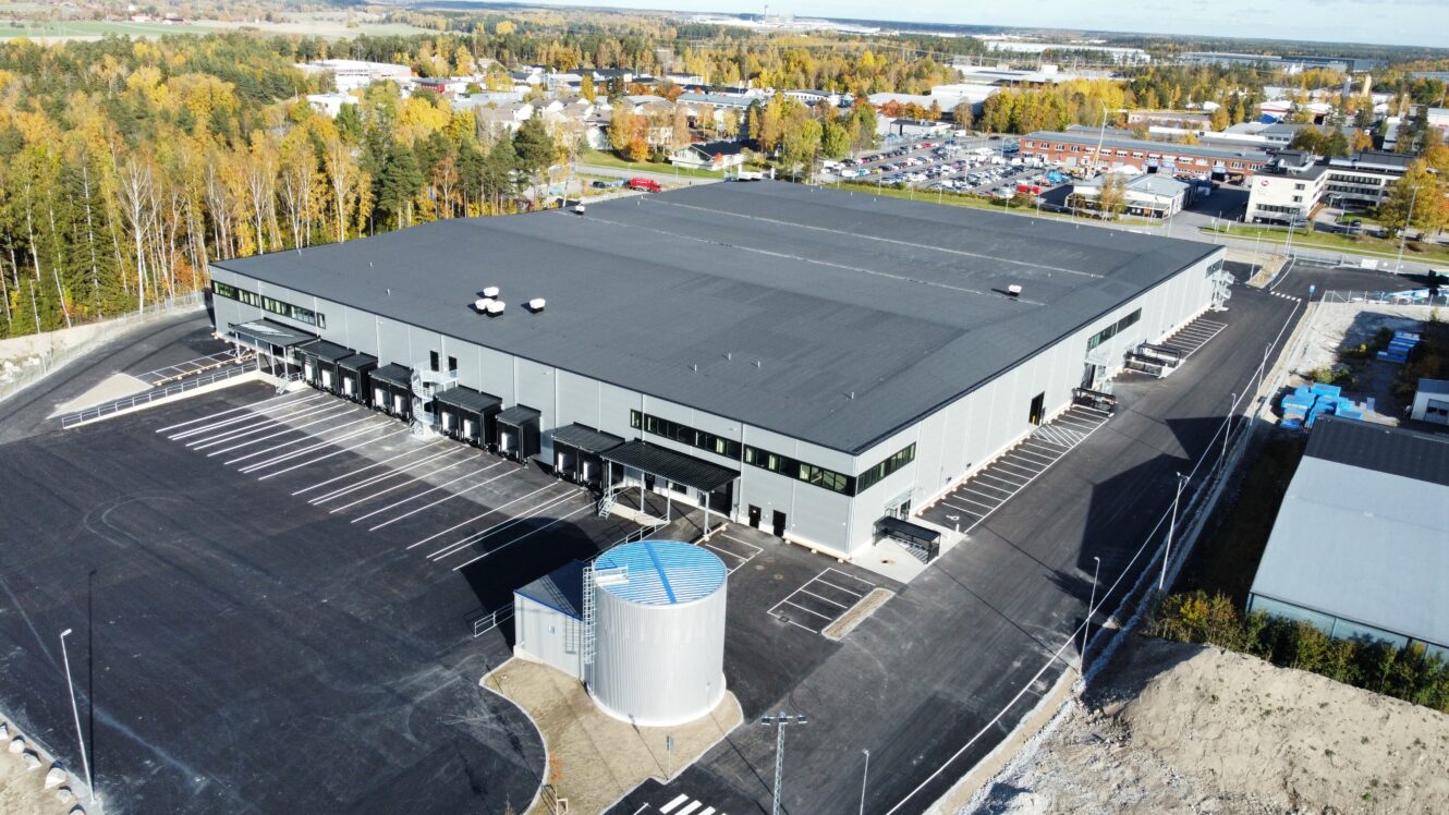 Ett stort industrilager med flera lastkajer och en rymlig parkeringsplats, omgiven av träd och andra byggnader en klar dag.