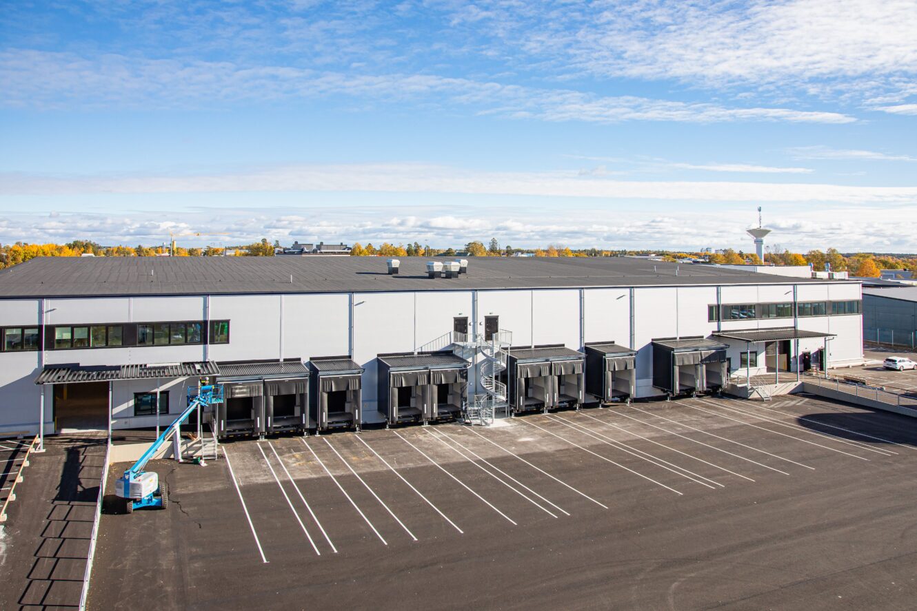 Stor modern lagerbyggnad med flera lastbryggor och en tom parkeringsplats framför, under en delvis molnig himmel.
