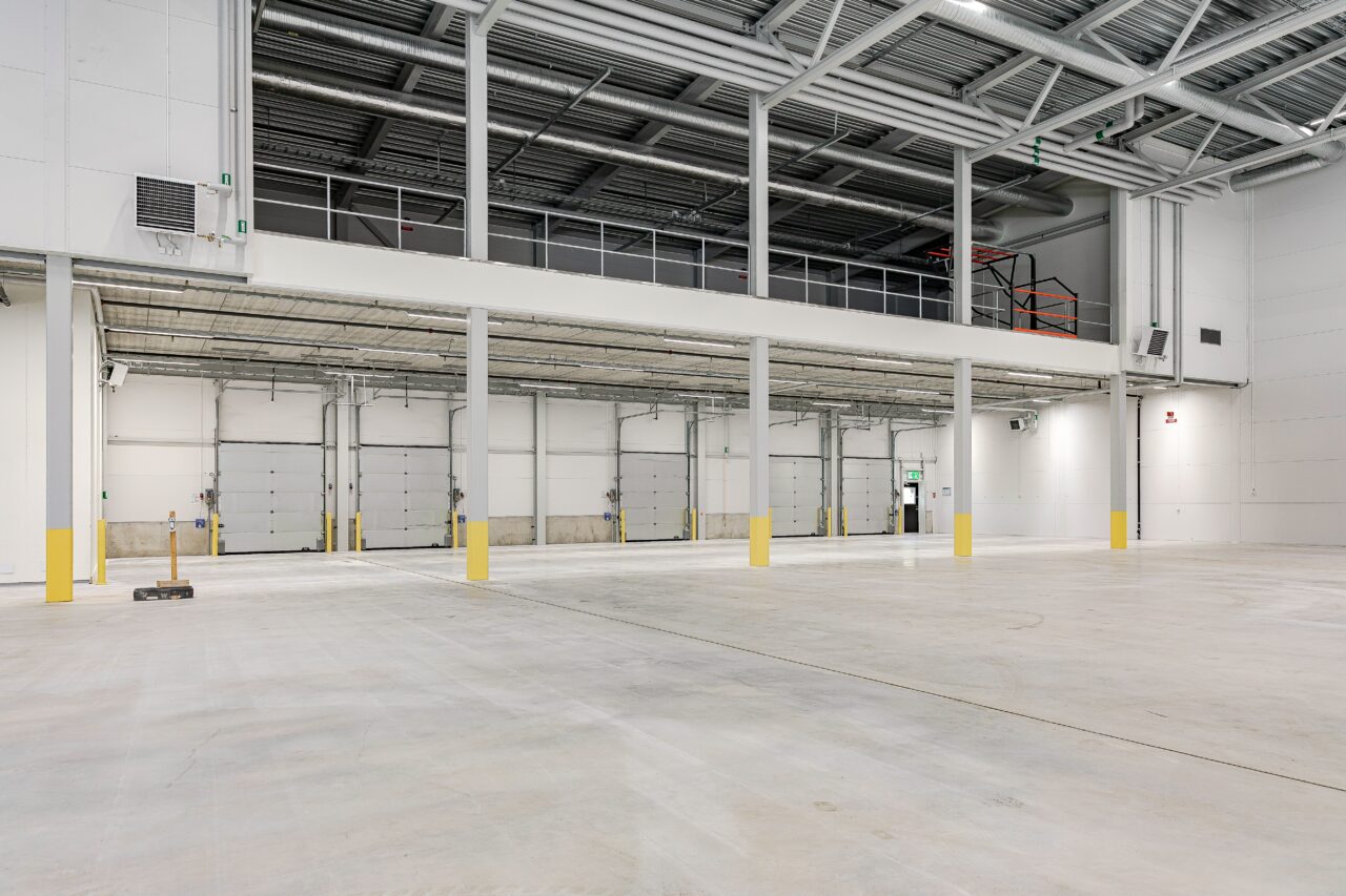 Stort, tomt industrilager med betonggolv, högt i tak, flera lastbryggor och en mezzaninvåning ovanför. Gula säkerhetsmarkeringar längs stödpelarna.