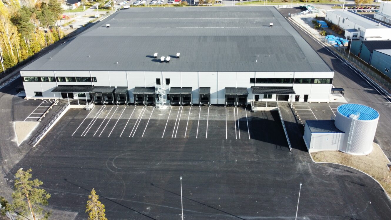 Flygfoto över ett stort, modernt industrilager med lastkajer, tomma parkeringsplatser och en intilliggande cylindrisk lagringstank.