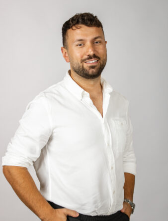 En man med kort lockigt hår och skägg, klädd i en vit skjorta med knappar, står med händerna i fickorna mot en ljus bakgrund.