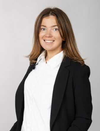 En kvinna med brunt hår i svart kavaj och vit skjorta ler mot kameran mot en ljus bakgrund.