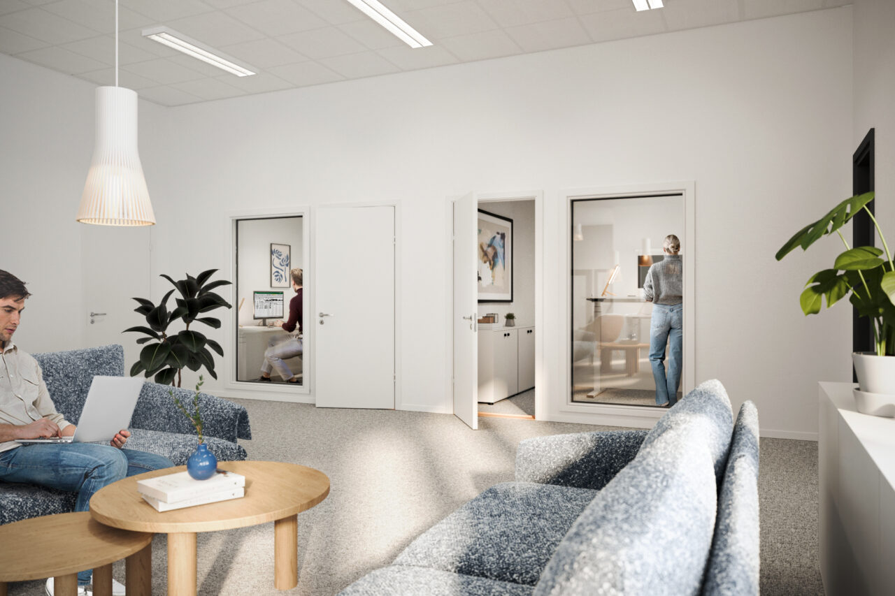 Modernt kontorsrum med blå soffor, ett runt träbord och krukväxter; två personer arbetar i glasrum i bakgrunden medan en annan använder en bärbar dator i förgrunden.