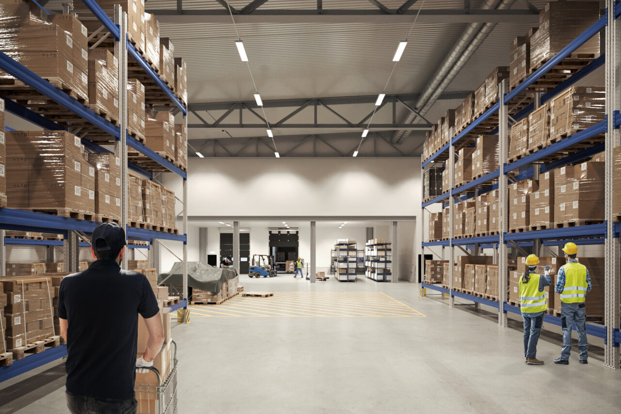 Rymlig lagerlokal med höga hyllor fulla av lådor; tre arbetare iförda skyddsvästar och hjälmar syns, och en person skjuter en vagn.