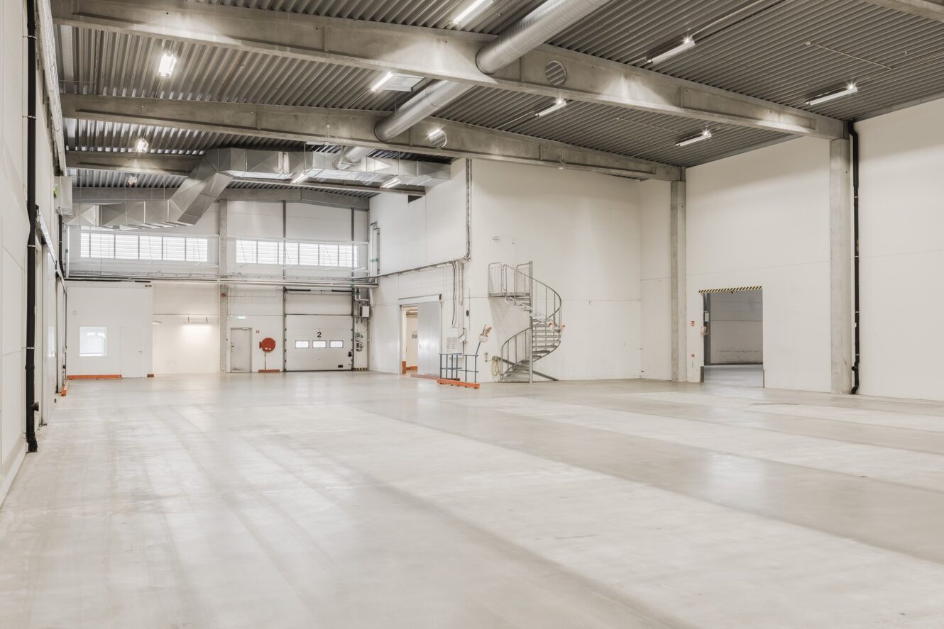 Rymlig, tom industrihall med högt i tak, betonggolv, vita väggar, stora dörrar och en spiraltrappa i metall nära bakväggen.