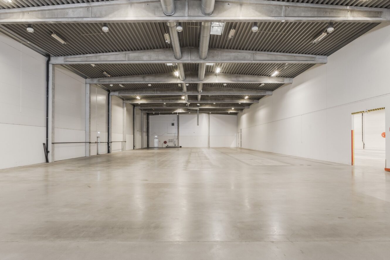 Tomt industriellt lagerutrymme med betonggolv, högt i tak, vita väggar, takbelysning och synliga rör.