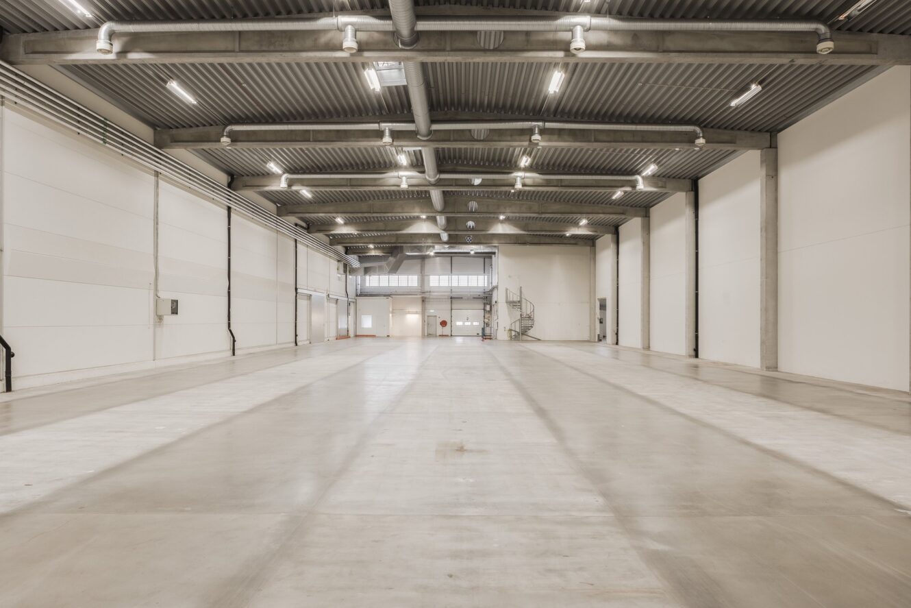 Stor tom lagerlokal med högt i tak, synliga bjälkar, stark takbelysning och släta betonggolv. Vita väggar avgränsar det rymliga öppna området.