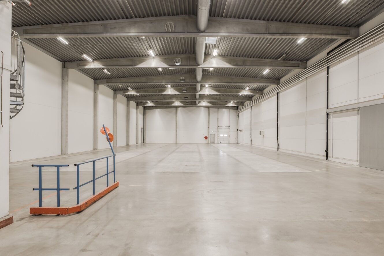 Stor tom lagerlokal med betonggolv, högt i tak, synliga bjälkar, vita väggar och minimalt med synlig utrustning.