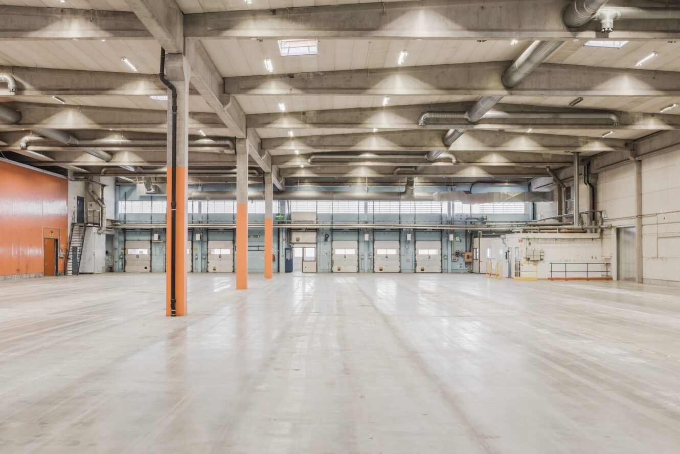 Rymlig, tom lagerlokal med betonggolv, högt i tak, synliga rör, orange pelare och stora lastkajsdörrar längs bakväggen.