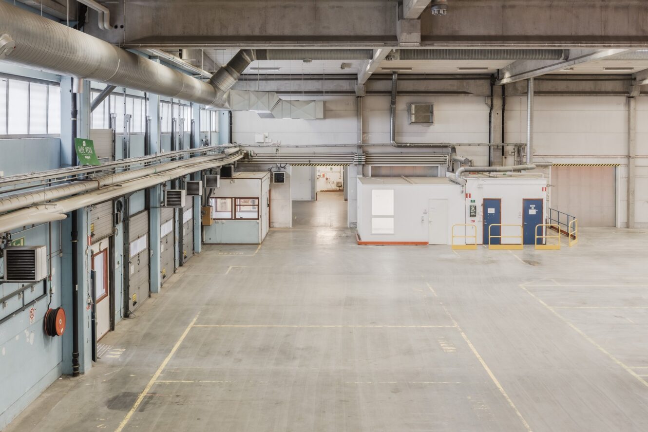 Stor, tom industrilagerlokal med betonggolv, högt i tak, lastkajer och två små vita kontor längs ena väggen.