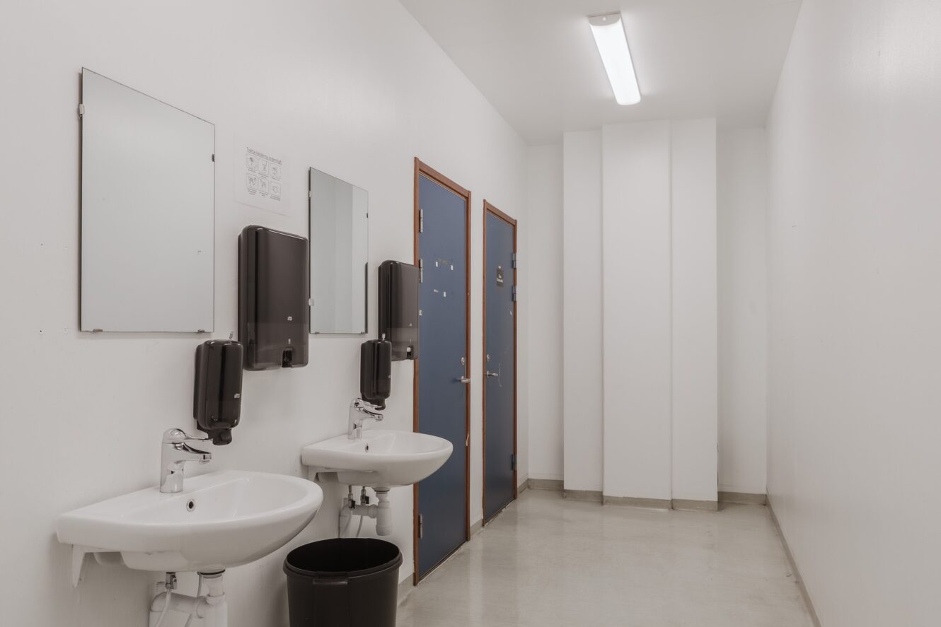 En ren, vit toalett med två handfat, två speglar, två tvålbehållare, en papperskorg och två stängda blå dörrar till båsen. Fluorescerande taklampa lyser upp utrymmet.