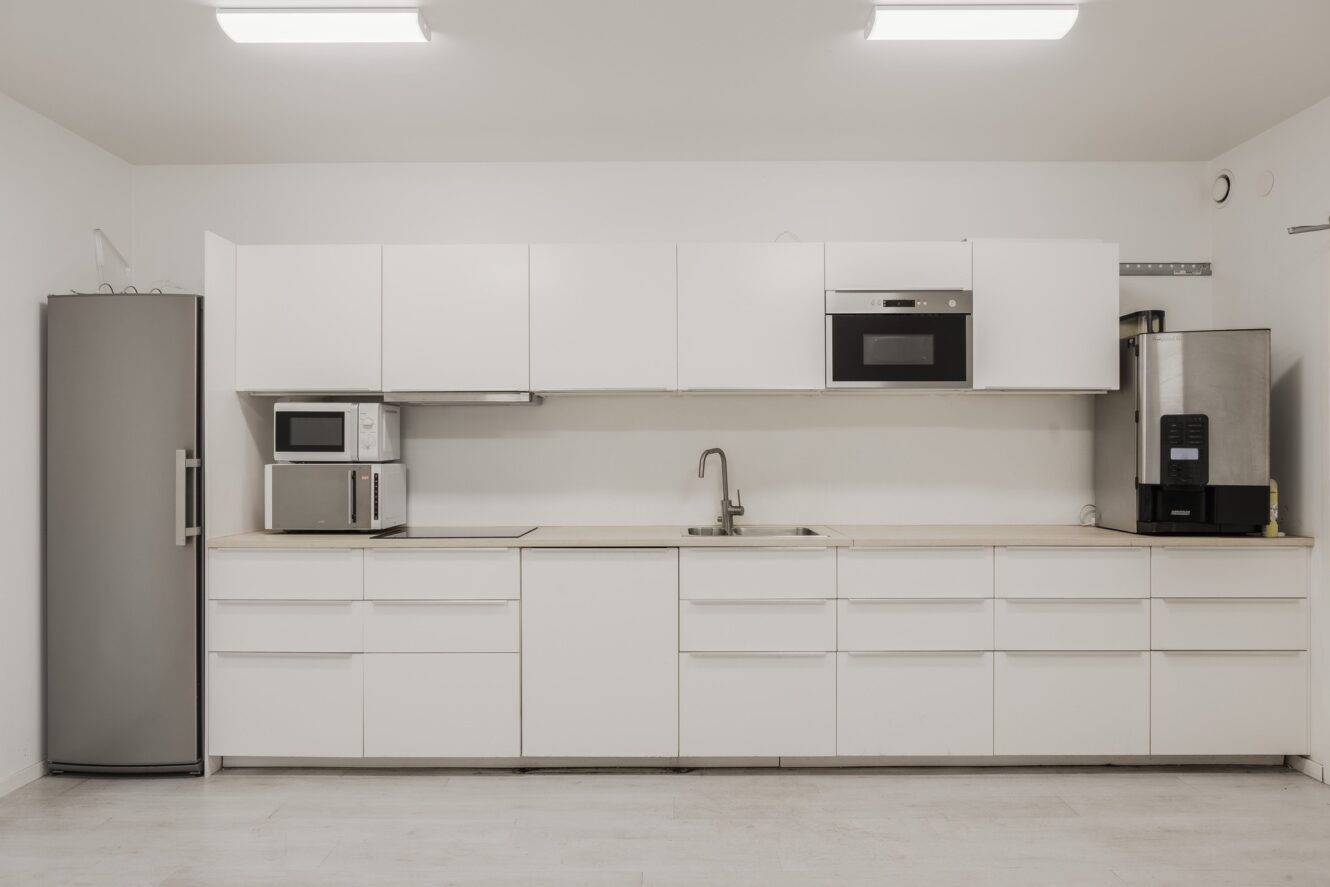 Minimalistiskt modernt kök med vita skåp, kylskåp, mikrovågsugn, ugn, kaffebryggare och diskho i rostfritt stål under starka taklampor.