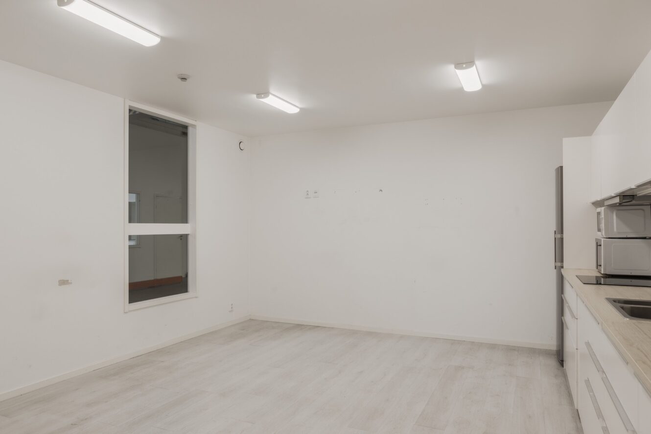 Ett i stort sett tomt, modernt kök med vita väggar, ljust trägolv, lysrör i taket, ett fönster och en rad köksmaskiner på höger sida.