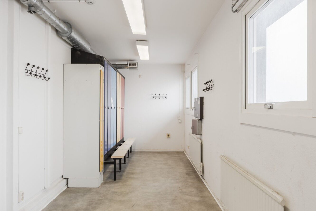Ett rent, tomt omklädningsrum med skåp, en bänk, klädkrokar på väggarna, fönster och en radiator längs golvet.