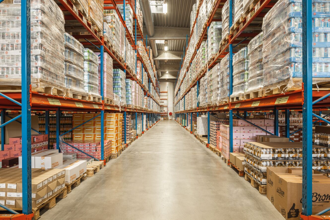 En lagergång kantad av höga hyllor staplade med pallar av olika varor och produkter i lådor, som sträckte sig till byggnadens bortre ände.