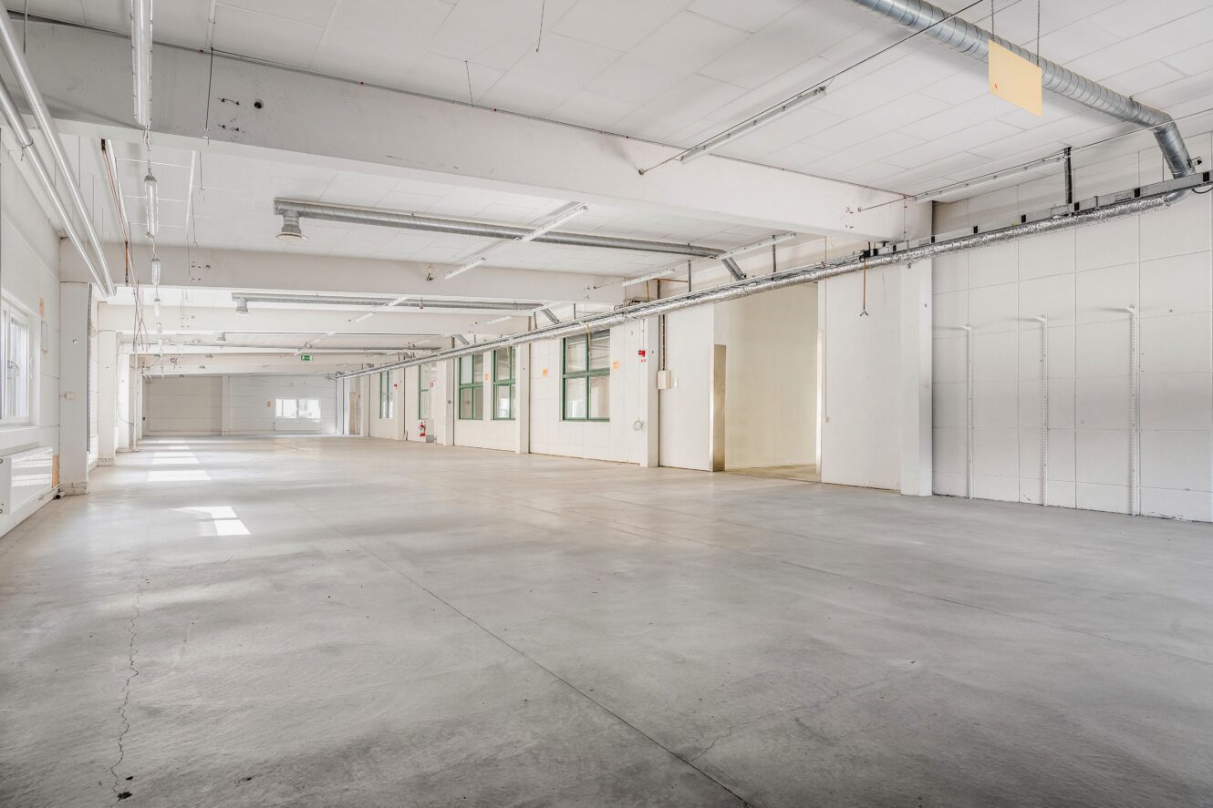 Stort tomt industrilager med betonggolv, vita väggar, rör i taket och fönster längs ena sidan, upplyst av naturligt och artificiellt ljus.