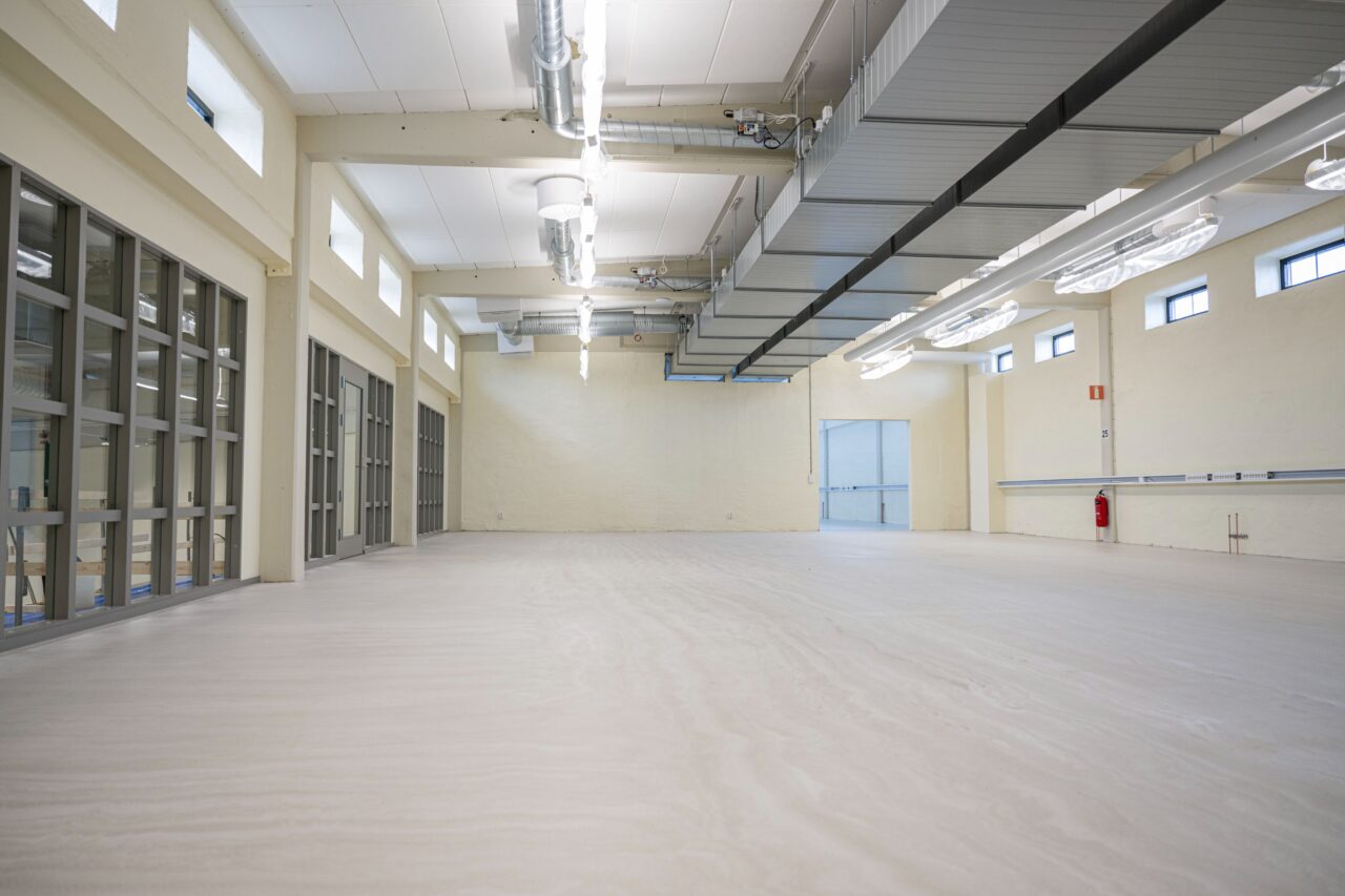 Rymlig, tom industrilokal med högt i tak, stora fönster, synliga ventilationskanaler och stark belysning. Krämfärgade väggar och ljust golv.
