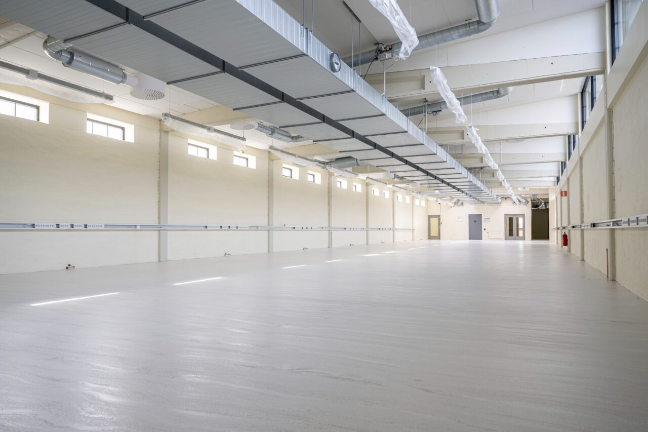 En stor, tom industri- eller handelslokal med vita golv, beige väggar, högt i tak, synliga kanaler och lysrör.