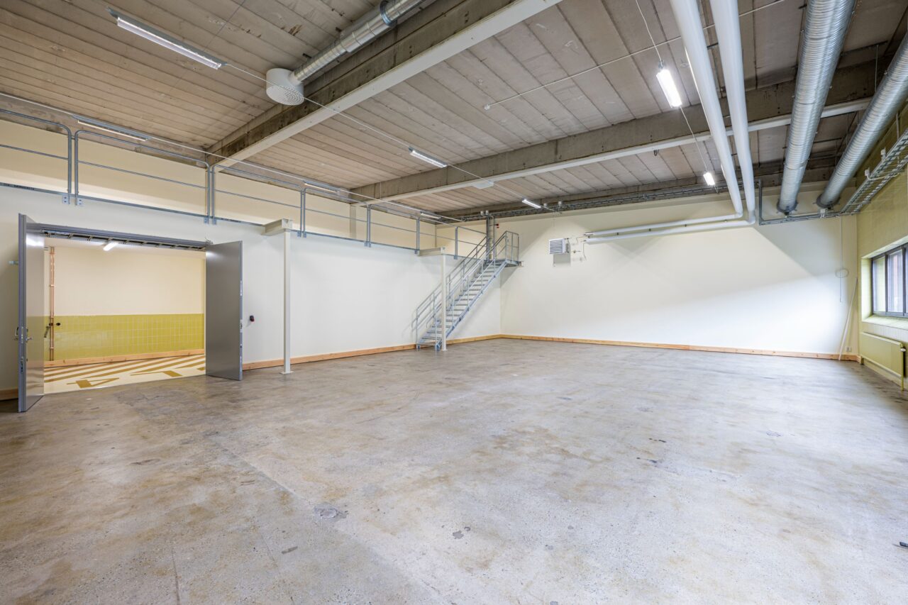 Rymlig, tom industrilokal med betonggolv, högt i tak, metalltrappa, stora fönster och synliga rör. Dubbeldörrar leder till ett kaklat område.