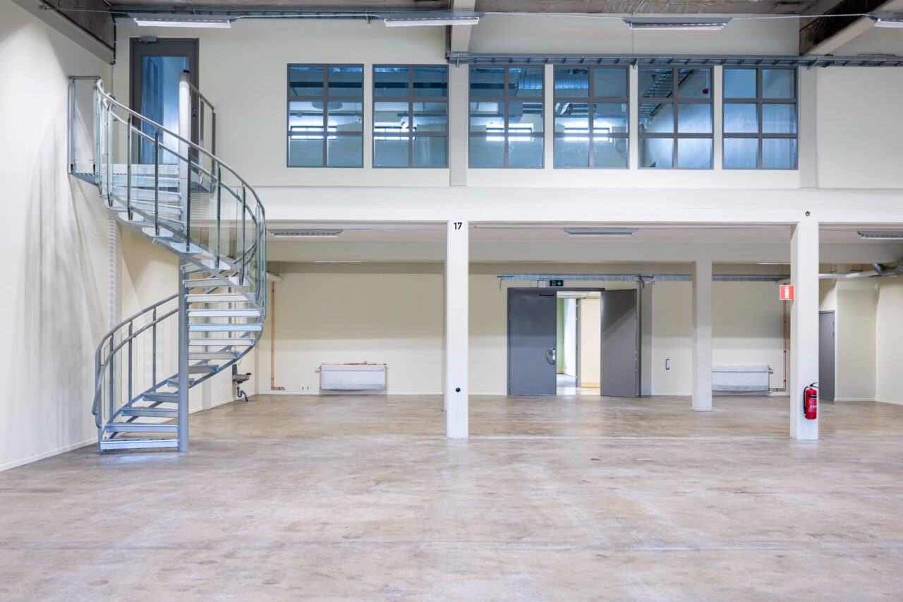 Interiör i en tom industribyggnad med en spiraltrappa i metall, stort öppet golv, högt i tak och en övre våning med glasfönster och dörrar.