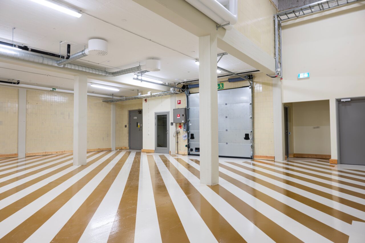 Rymlig industrilokal med brun- och vitrandigt golv, beige kaklade väggar, ståldörrar, rör i taket och synliga nödutgångsskyltar.