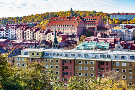 En stadsbild som visar färgglada bostadshus med röda tak och en kulle med träd och ett torn i bakgrunden under en delvis molnig himmel.