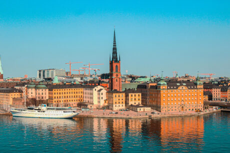 Panoramautsikt över Stockholms hamn med historiska byggnader, en kyrka med hög spira, ett vitt fartyg i hamn och byggkranar i bakgrunden under en klarblå himmel.