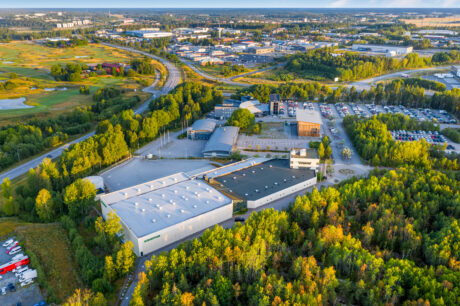 Flygfoto över en företagspark omgiven av träd, parkeringsplatser och vägar, med en golfbana och en stadsbild i bakgrunden.
