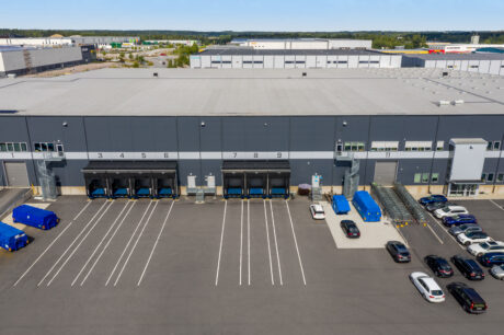 Flygfoto över ett stort industrilager med flera lastbryggor, parkerade bilar, blåa containrar och tomma parkeringsplatser framför.