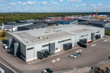 Flygfoto över ett stort industrilager med lastkajer, parkerade bilar och omgivande kommersiella byggnader under en blå himmel.