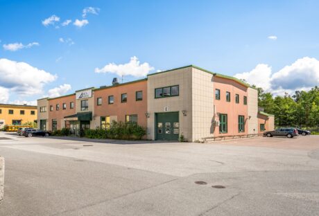 En affärsbyggnad i två våningar med beige och orange fasad, omgiven av en parkeringsplats med några parkerade bilar, mot en blå himmel med spridda moln.