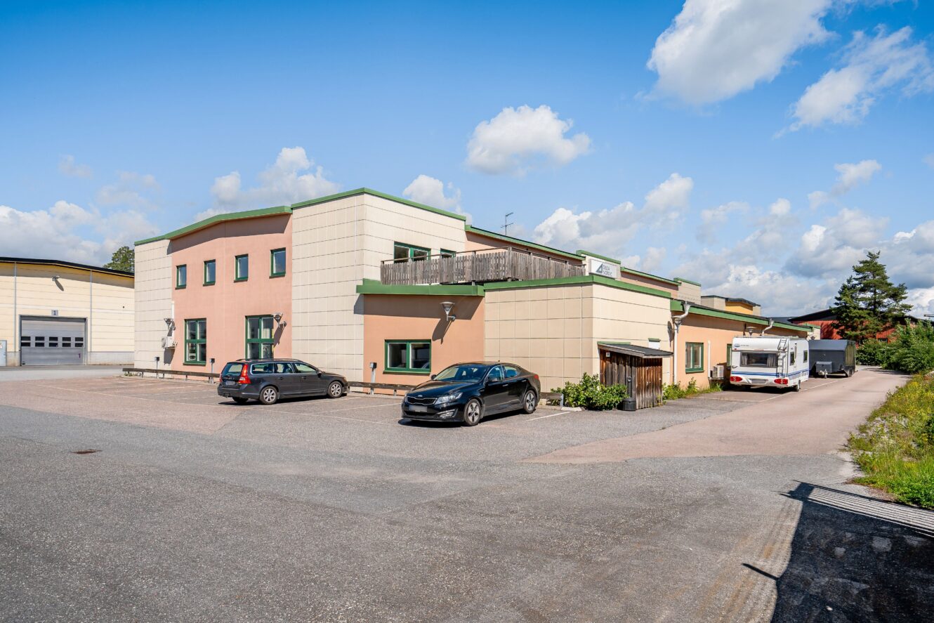 En beige affärsbyggnad med grön dekor, bilar parkerade framför och en husvagn vid sidan om på en asfalterad tomt under en delvis molnig himmel.