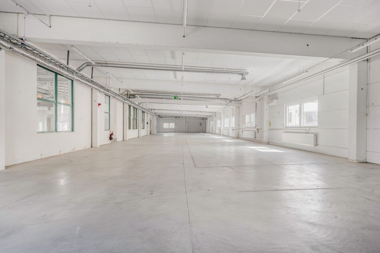 Stort, tomt industrilager med vita väggar, högt i tak, synliga rör och flera fönster som släpper in naturligt ljus i den rymliga interiören.