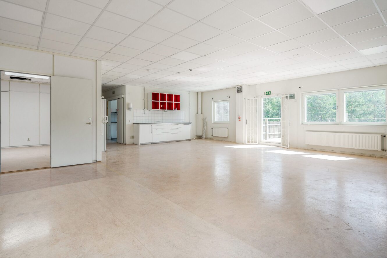 Rymligt, tomt rum med vita väggar, kaklat tak, stora fönster, en köksdel med röda hyllor och ett beige golv. Utrymmet fylls av naturligt ljus.