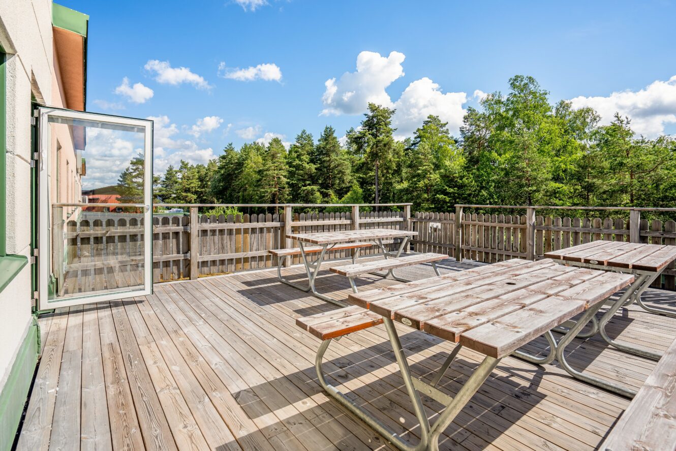 Trädäckad uteplats med tre picknickbord, omgiven av ett trästaket, mot ett skogsområde under en solig, delvis molnig himmel. En öppen glasdörr leder ut.