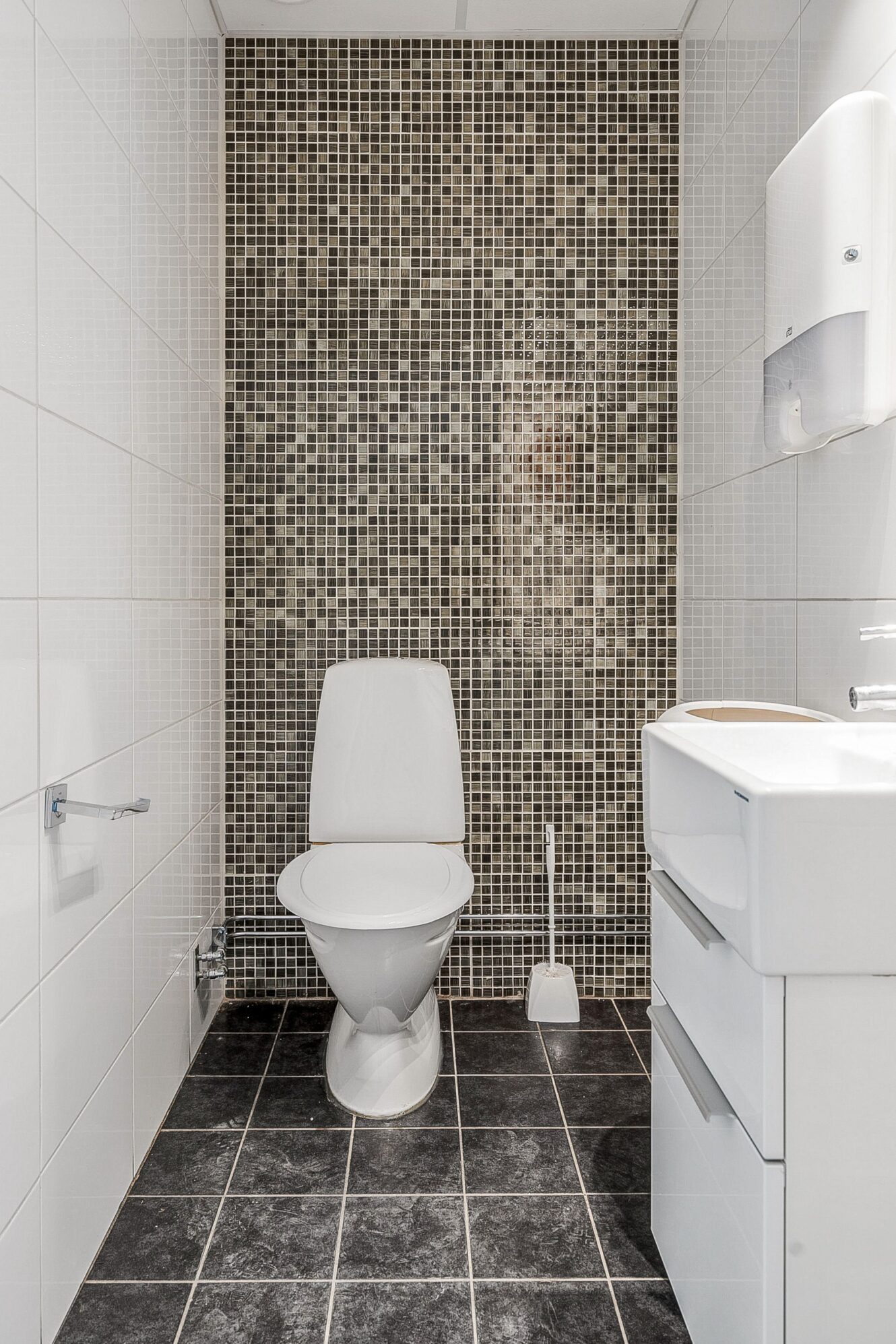 Ett modernt badrum med en vit toalett, väggmonterat handfat, toalettborste, pappersdispenser och mörka mosaikplattor på bakväggen.