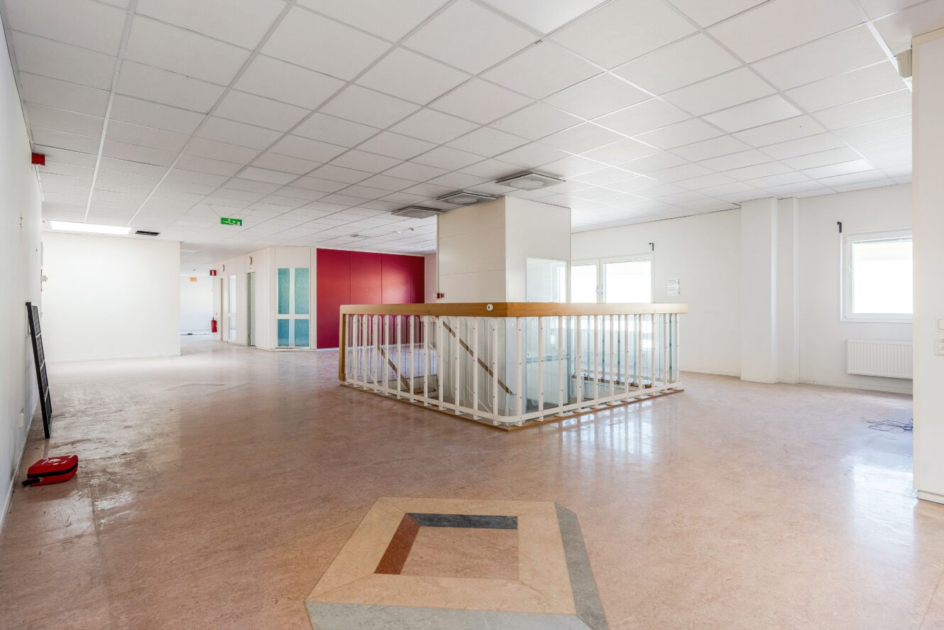 En rymlig, tom interiör med tegeltak, stora fönster och en central trappa som omges av ett vitt räcke. Golvet är ljust och en vägg är rödmålad.