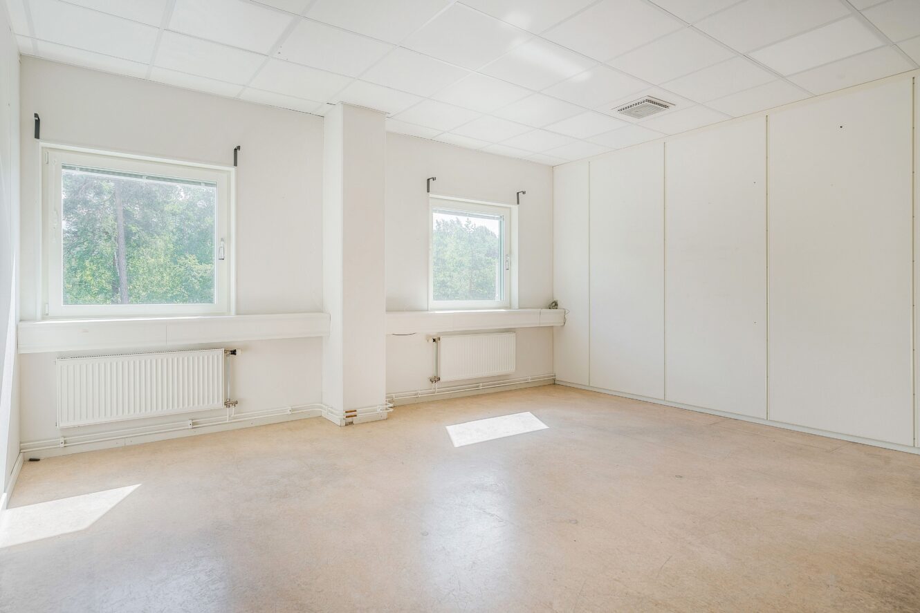 Tomt, omöblerat rum med ljusa väggar, två fönster som släpper in dagsljus, beige golv och vitmålat tak. Radiatorer är installerade under båda fönstren.