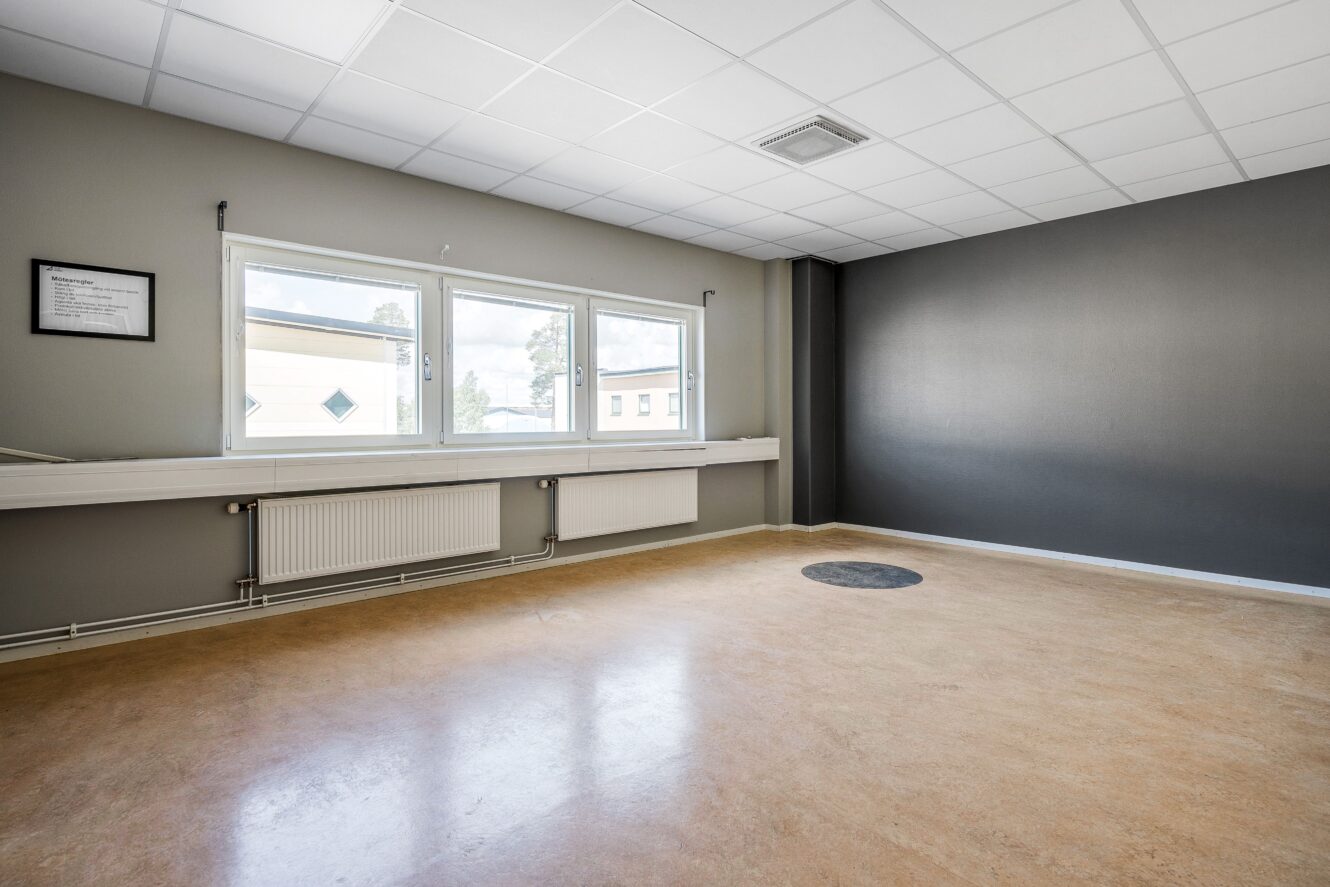 Tomt rum med ljusbrunt golv, grå väggar, tre stora fönster, ett väggradiator och ett inramat föremål på väggen. Taket har en ventil och infälld belysning.